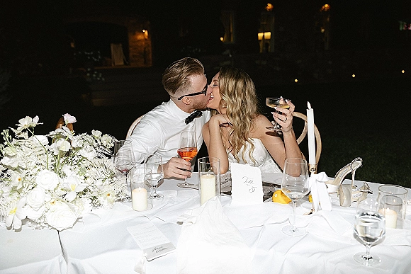 Wedding kiss at the sweetheart table, bride holding a champagne coupe as taper candlelight glows in a dim reception venue