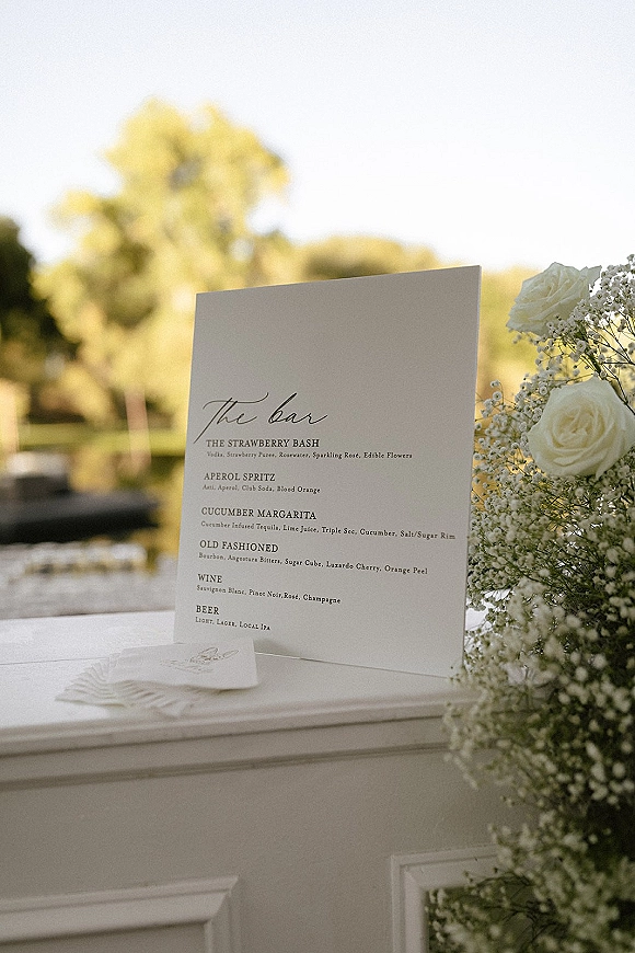 Bar menu sign with wedding bar menu details on a printed card, framed by white roses and baby’s breath on a bar counter outdoors