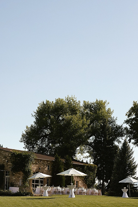 Outdoor reception setup with cocktail tables and white tablecloths under patio umbrellas, wooden chairs and string lights by a stone building lawn