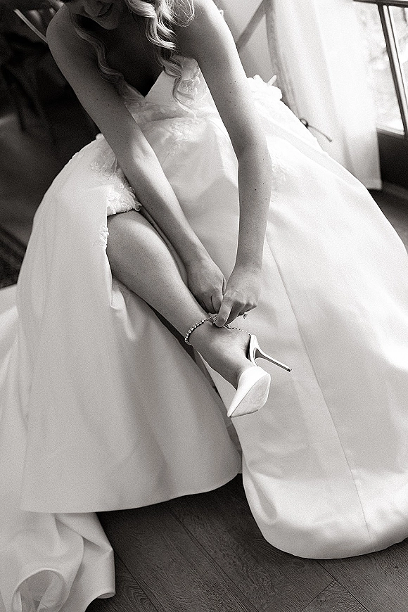 Bride putting on shoes in window light, showing white pointed toe heels and an ankle bracelet beside her wedding dress on hardwood floor