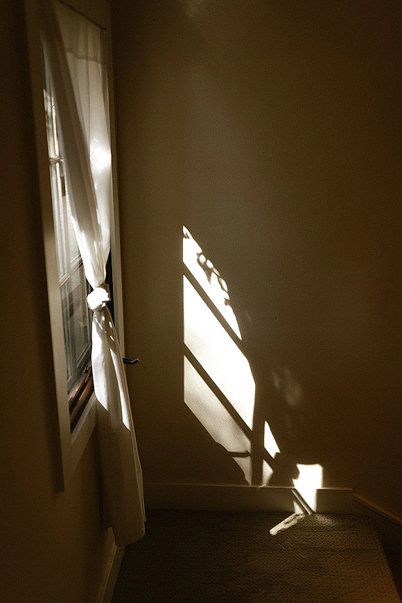 Bridal robe hanging by a window with soft sunlight casting shadows on the interior wall, with white curtains and a window handle nearby