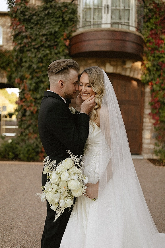 Couple portrait of bride and groom embrace as he kisses her cheek, holding a white rose bouquet before an ivy-covered stone building door