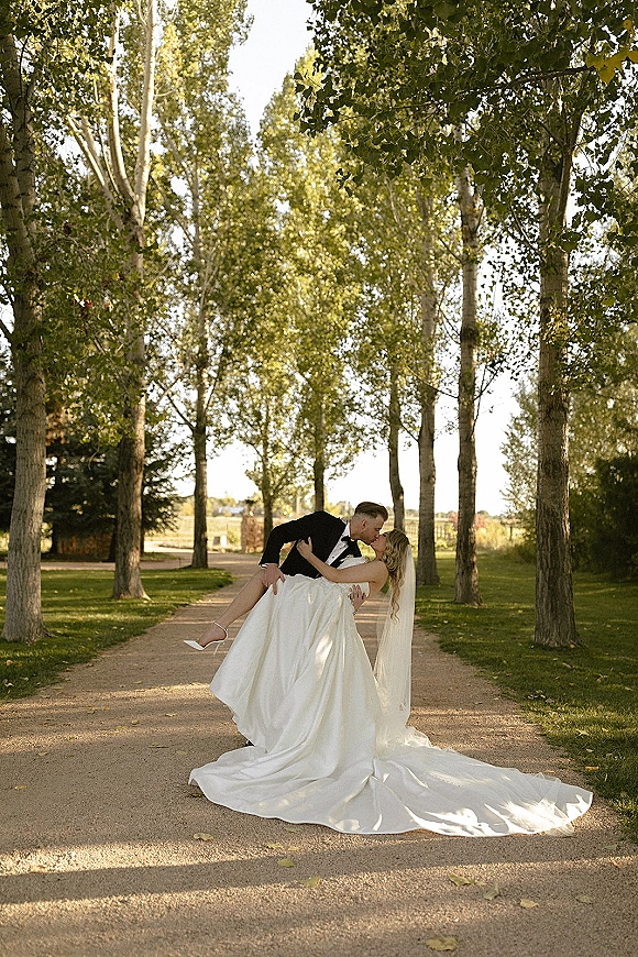 Wedding kiss portrait of groom lifting bride into a dip kiss, her long veil and dress train flowing along a sunlit tree-lined path