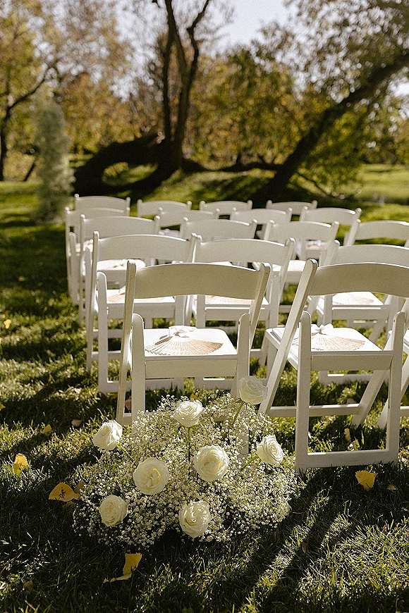 Outdoor ceremony seating with white folding chairs and paper fans, lined along a grassy aisle with white rose and baby's breath flowers in sunlight