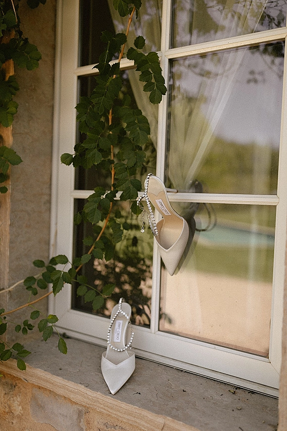 Wedding shoes, bridal heels in white pointed-toe style with a crystal strap resting on a window ledge beside sheer curtains and vines