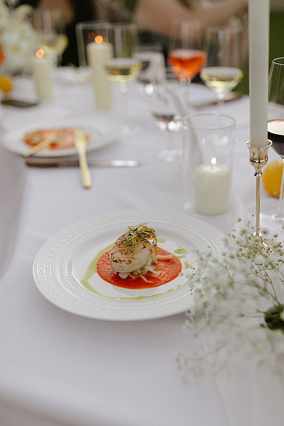 Wedding tablescape with white wedding tablescape details, gold flatware, wine glasses, baby's breath, candles, and plated appetizer amid outdoor greenery