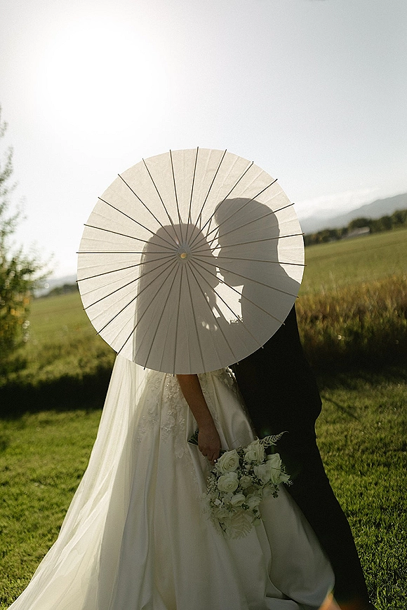 Wedding kiss portrait of bride and groom kissing in a sunlit field, bride holding a white paper parasol and bouquet, hills behind