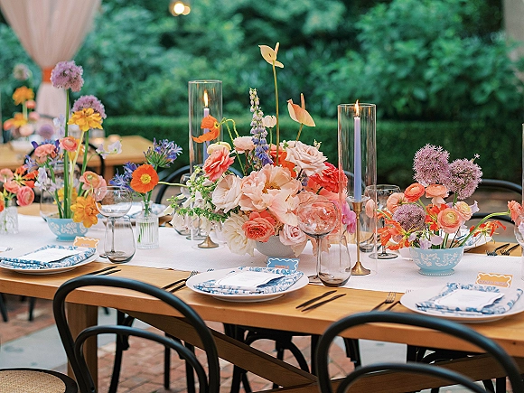 Reception tablescape with colorful wedding tablescape florals, lavender taper candles, blue patterned plates, and menus on a wood table under bistro lights.