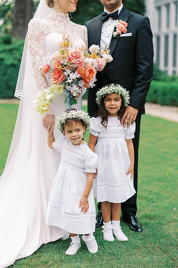 Wedding family portrait of bride and groom with flower girls, bride in lace high-neck gown and veil holding peach-coral bouquet on garden lawn