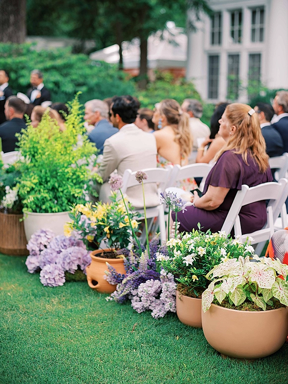 Ceremony guests seated in white folding chairs at a garden wedding ceremony, with potted plants and purple florals along the aisle edge