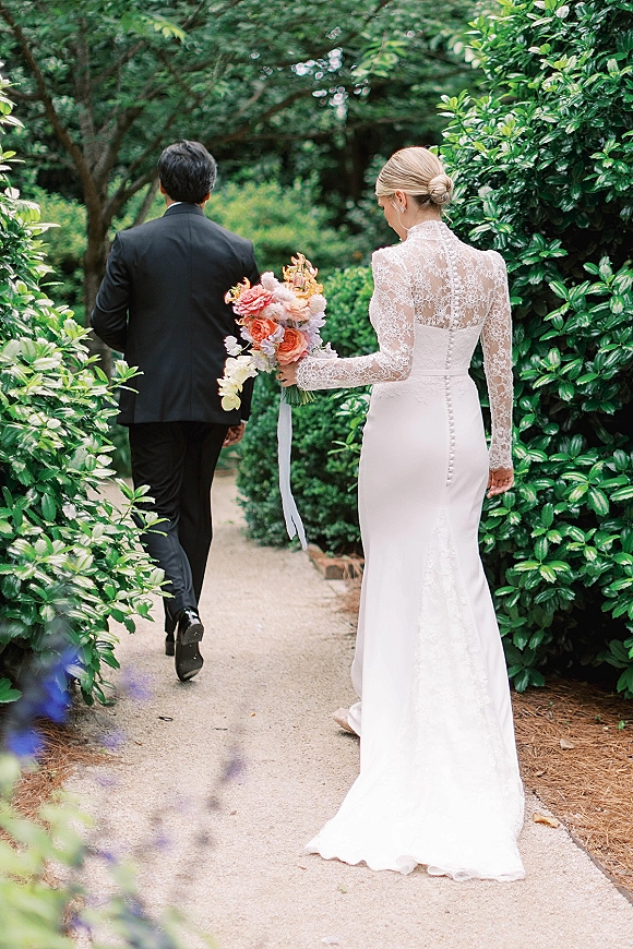 First look moment as bride approaches groom from behind on a garden path, holding a colorful bouquet with a long ribbon