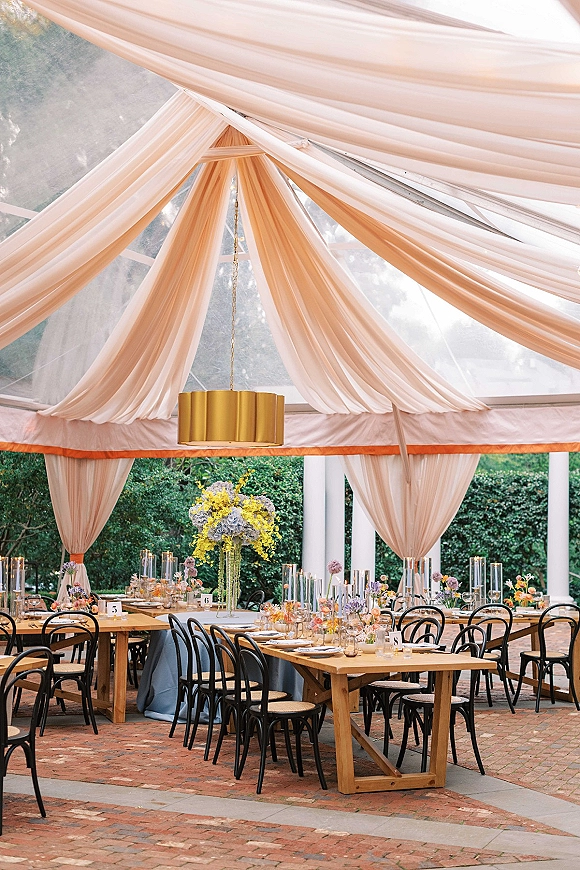 Reception tablescape under a clear top tent with draped fabric ceiling, chandelier, long wooden tables, candles, and floral centerpieces in a garden patio setting