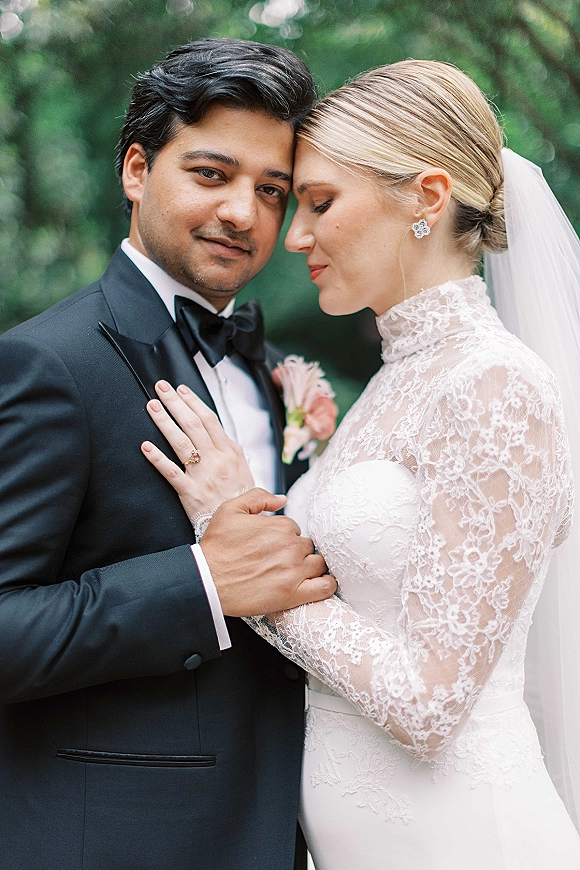 Couple portrait of bride and groom close up, touching foreheads; her lace long-sleeve veil and his black tuxedo against garden foliage