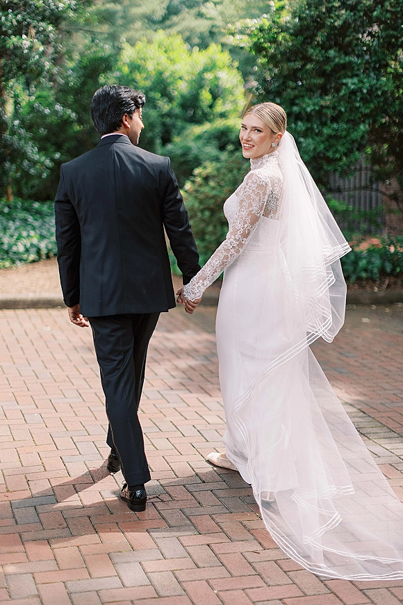 Couple portrait of bride and groom walking away holding hands, her cathedral veil flowing over a brick garden path with trees behind