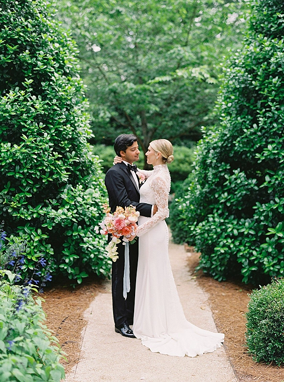 Couple portrait of bride and groom embrace on a garden path, bride holding a colorful bouquet with long ribbon, lace sleeves and dress train