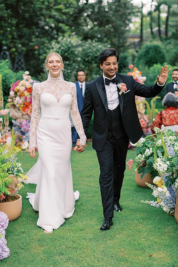 Wedding recessional as bride and groom walk hand in hand down a floral-lined garden aisle, bride in lace long sleeves and veil waving to guests
