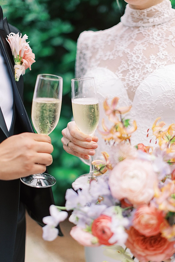 Wedding toast with bride and groom clinking champagne flutes, bouquet and boutonniere details against soft green foliage backdrop