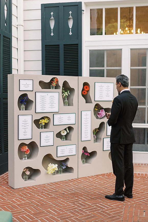 Seating chart display with wedding seating chart ideas, framed escort cards in wall niches with bud vase flowers on a brick patio backdrop