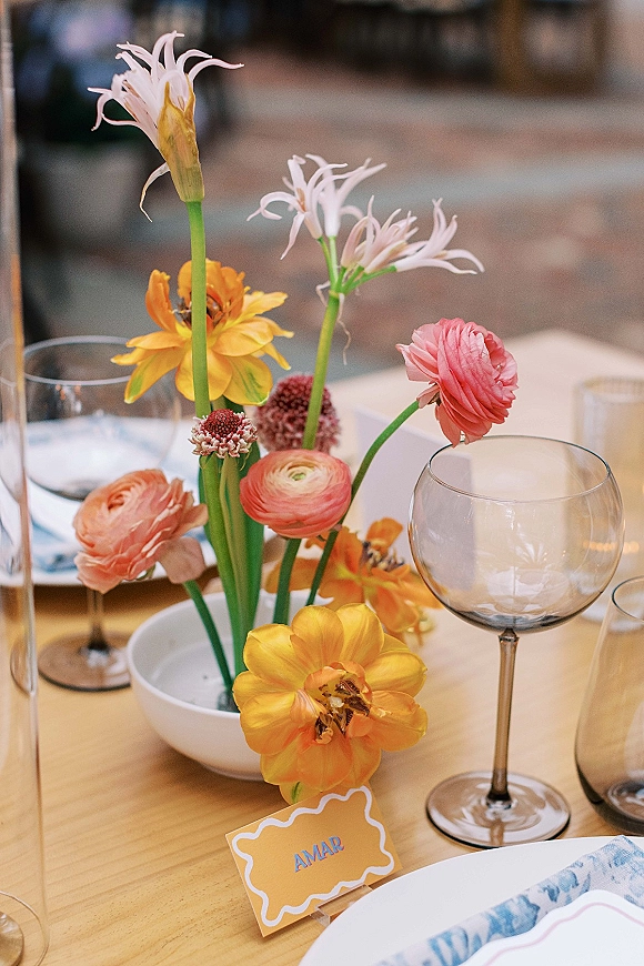 Reception tablescape with colorful wedding centerpiece of ranunculus and tulips, glass goblets and votives, and place cards on a wood table