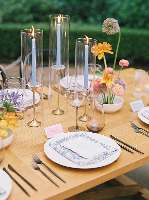 Reception tablescape with taper candle centerpiece, brass candlesticks, glass hurricanes, bud vase flowers, and place cards on a wooden table outdoors