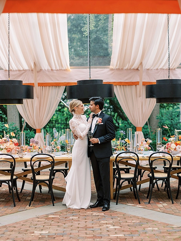 Couple portrait of bride and groom champagne toast, holding flutes under a tented garden reception with a long banquet table behind them