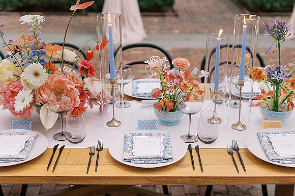 Reception tablescape with wedding table centerpiece of colorful wedding flowers, blue taper candles, and mixed glassware on a brick patio table