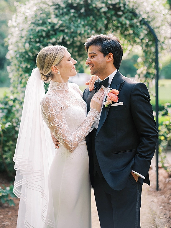 Couple portrait of bride adjusting groom’s black bow tie, lace long-sleeve dress and veil, forehead to forehead beneath a flowering arch outdoors