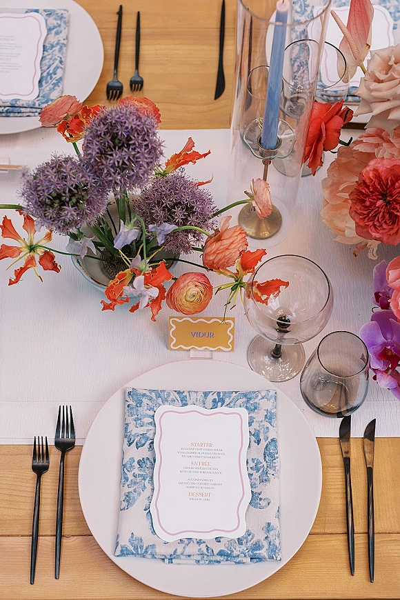 Wedding tablescape with wedding place setting, blue patterned napkins, menu cards, black flatware, wine glasses, and floral centerpiece on wood table