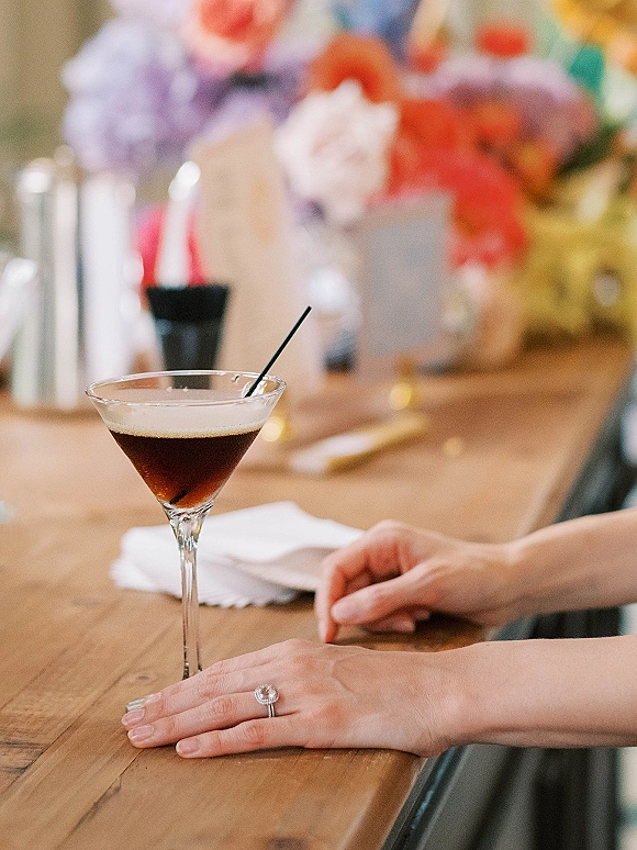 Wedding cocktail and signature wedding drink in a martini glass with straw, bridal hand showing engagement ring on a wood bar top