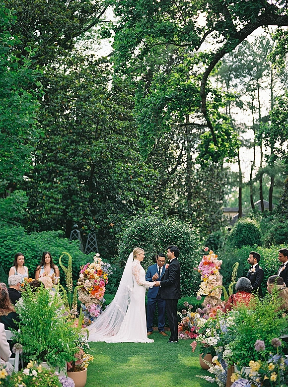 Wedding vows during an outdoor garden ceremony as bride and groom hold hands under colorful floral arch with bridal veil and wedding party nearby