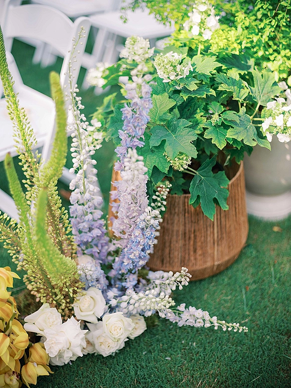 Ceremony aisle flowers in a woven basket with white roses, purple delphinium, and yellow blooms beside white folding chairs on a lawn