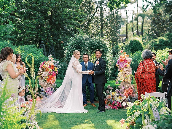 Wedding ceremony moment in an outdoor garden ceremony with bride and groom holding hands, long veil flowing as guests applaud by floral pillars