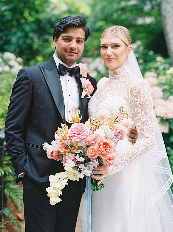 Couple portrait of bride in lace veil holding a blush bouquet beside groom in black tuxedo, posed amid soft garden greenery