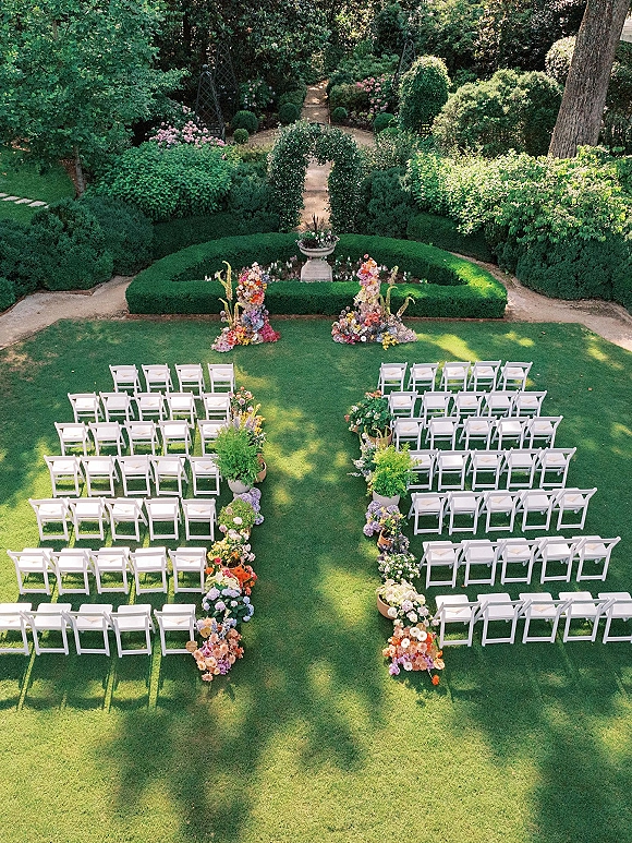 Outdoor ceremony setup with white folding chairs lining a stone path, colorful aisle flowers and potted plants on a manicured garden lawn