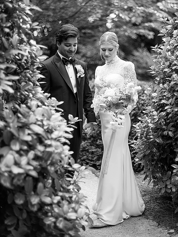 Couple portrait in a black and white wedding portrait style, bride in lace long sleeves holding a ribbon-wrapped bouquet, walking a garden path