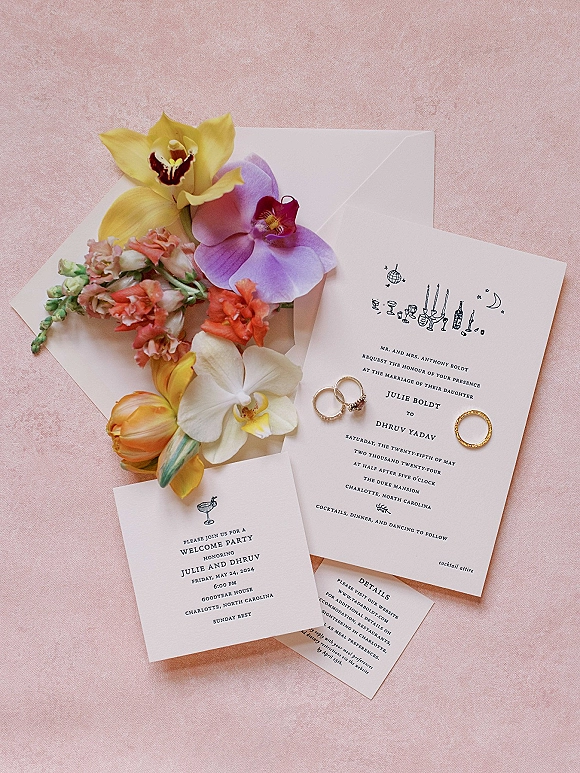 Invitation suite flat lay with envelopes, wedding invitation cards, orchids and tulips, plus gold rings arranged on a pink fabric backdrop