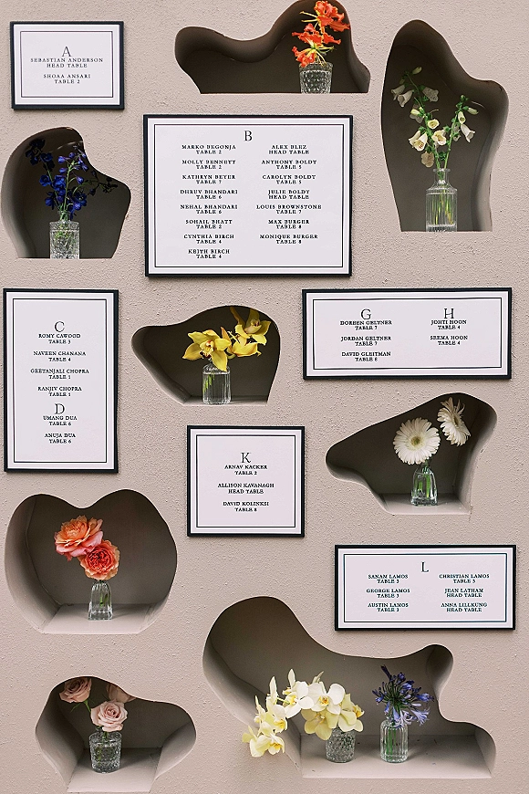 Seating chart display with black-framed wedding seating chart cards and glass bud vases of assorted flowers on a textured wall with niches
