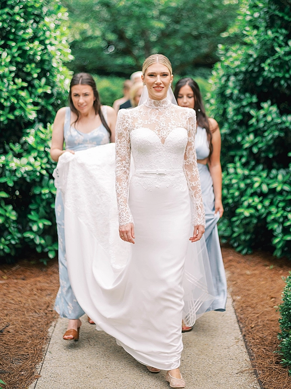 Bride portrait in a long sleeve lace wedding dress and veil, walking a garden walkway as bridesmaids lift her train beside hedges