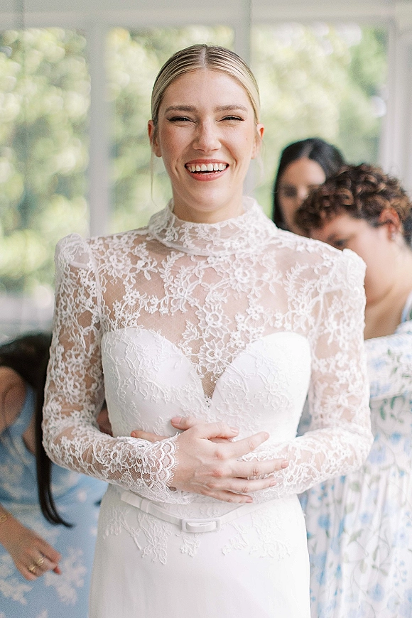 Bride portrait smiling in a high neck lace wedding dress with long sleeves and low bun, lit by window light in an indoor room