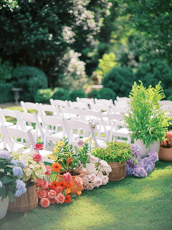 Ceremony seating with white folding chairs lined beside hydrangea and rose aisle florals in terracotta pots on a sunlit garden lawn