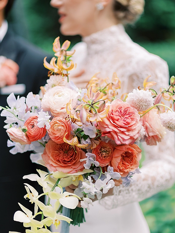 Bridal bouquet, garden rose bouquet of ranunculus, roses, orchids and sweet peas held by bride in high-neck lace dress outdoors with groom behind