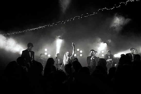 Wedding band performance with singer at microphones under stage lights and haze, string lights glowing above a packed dance floor crowd