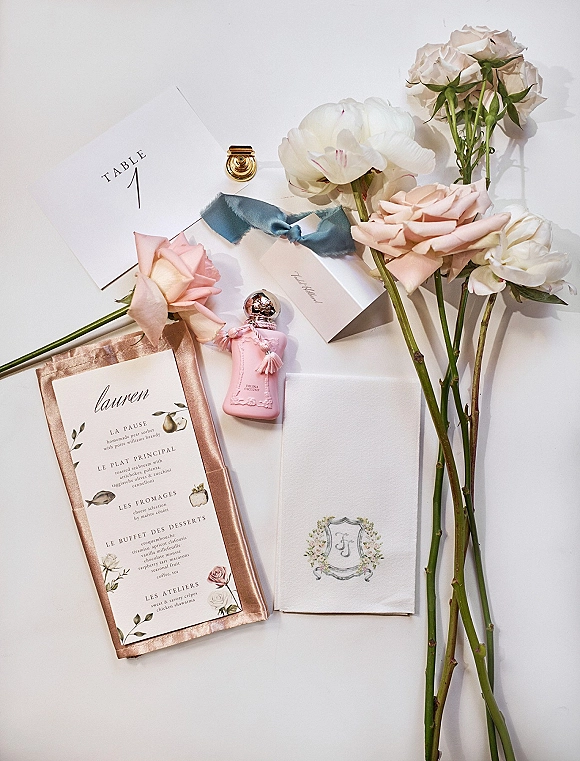 Wedding stationery flat lay with invitation suite, menu and table number card, blush florals, wax seal and silk ribbon on white surface