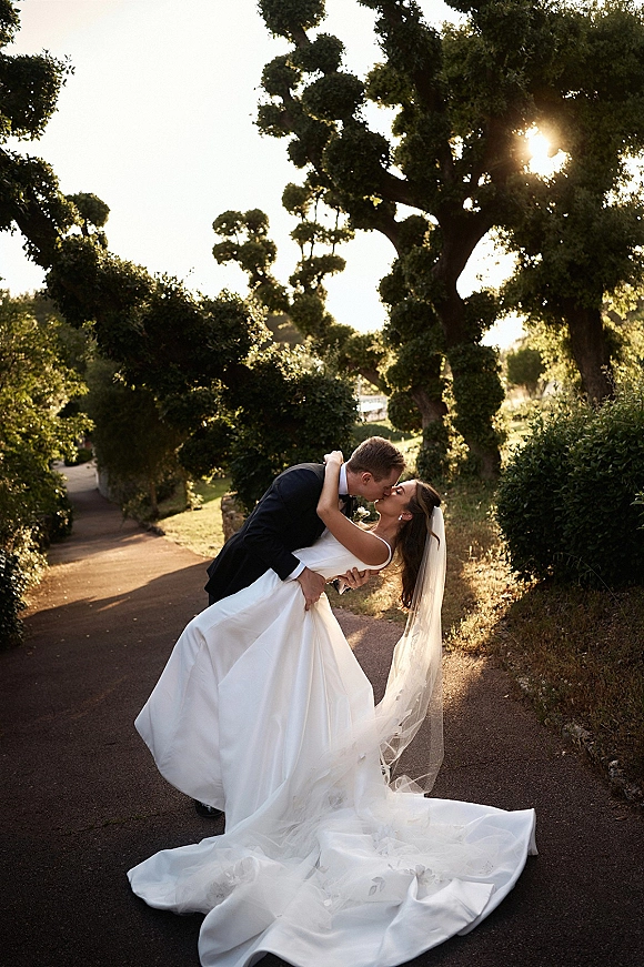 Wedding kiss portrait of bride and groom in a dip, her long veil and dress train flowing as sunlight glows on a tree-lined path