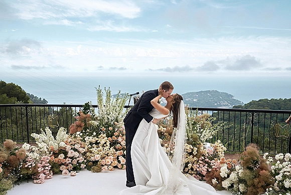 Wedding kiss portrait of bride dipped by groom, veil flowing over rose florals on a terrace balcony with ocean coastline view