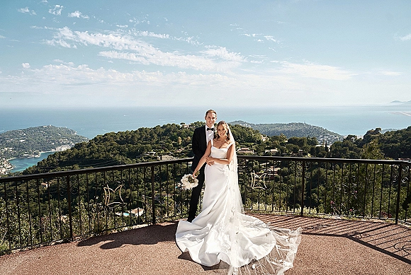 Couple portrait of bride and groom outdoors embracing, bride with bouquet and veil, by wrought-iron terrace railing with ocean view
