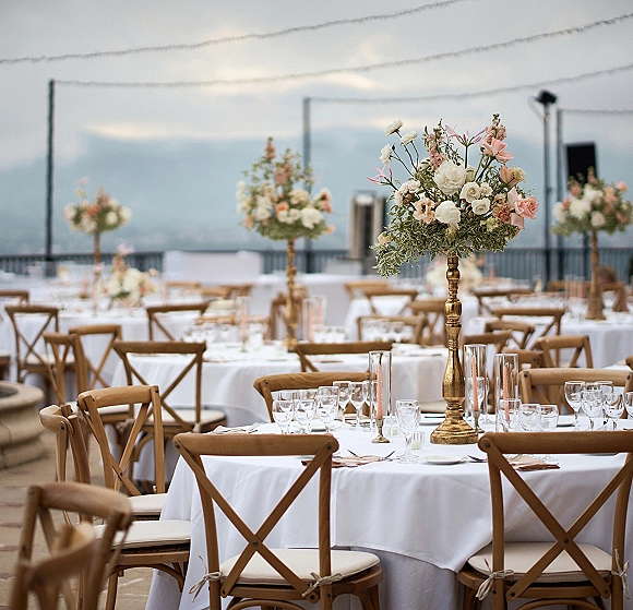 Reception tablescape with round wedding reception tables, tall blush and white floral centerpieces in gold vases on an outdoor terrace under string lights