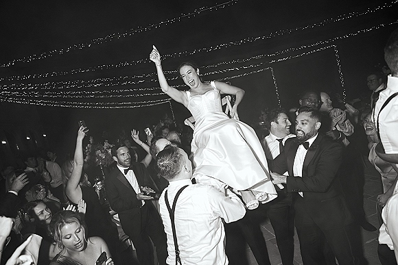 Wedding reception dancing as groomsmen lift the bride on a chair during the hora, with string lights over an outdoor patio crowd at night