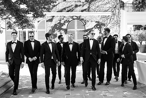 Groomsmen portrait of a group in black tuxedos and sunglasses, standing in a stone courtyard by a white ivy-covered facade under tree shade