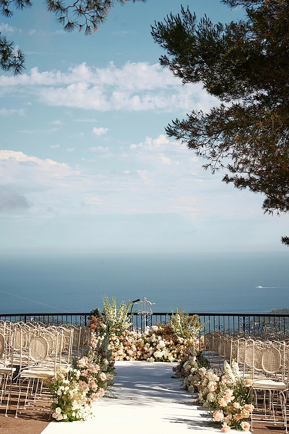 Ceremony aisle decor with a white aisle runner and pastel floral arrangements leading to wrought iron chairs on a terrace with ocean view